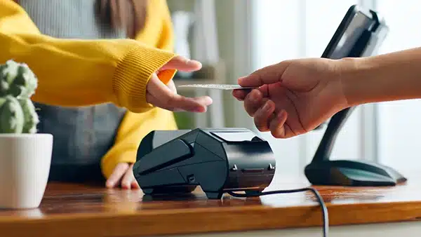 A close-up, realistic photo of a customer handing a debit card to a merchant at a wooden checkout counter with a modern payment terminal in a brightly lit shop
