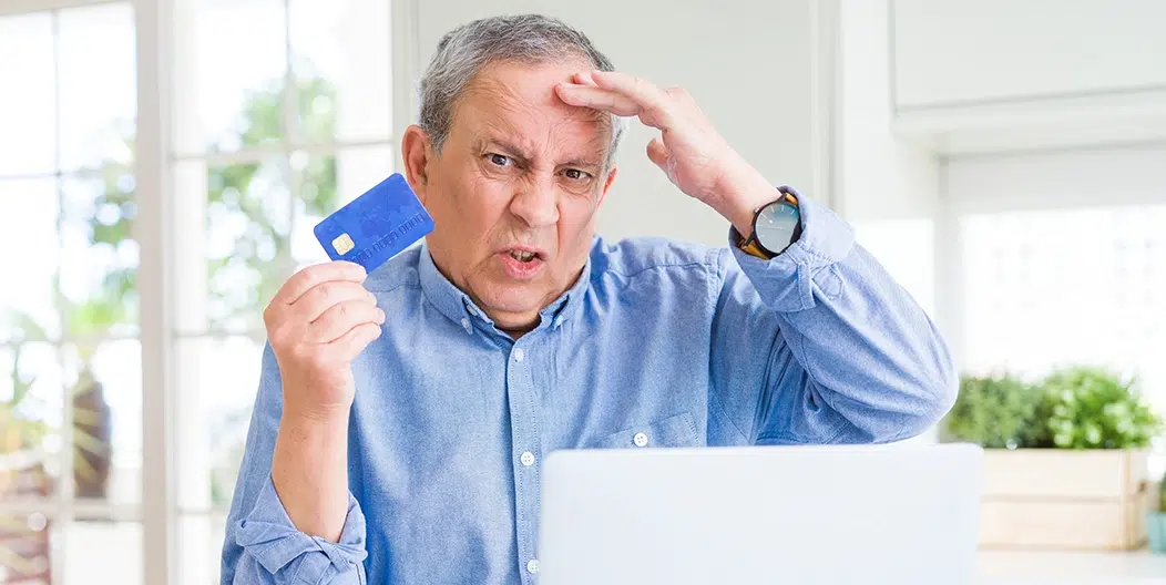 Older man looking frustrated while holding a credit card in front of a laptop, representing a declined payment during online checkout